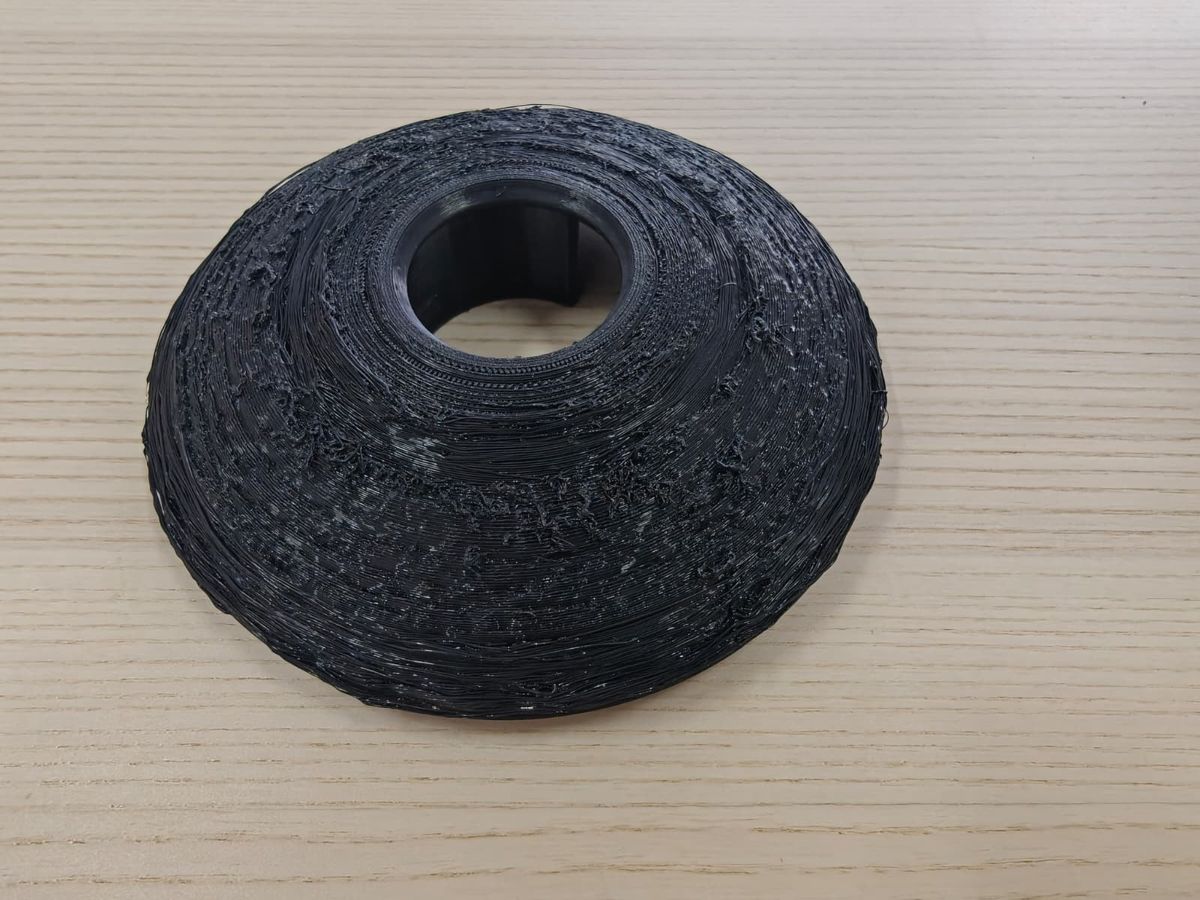 A black 3D printed cone-shaped part with loose strands and rough, stringy surfaces across its sloped face